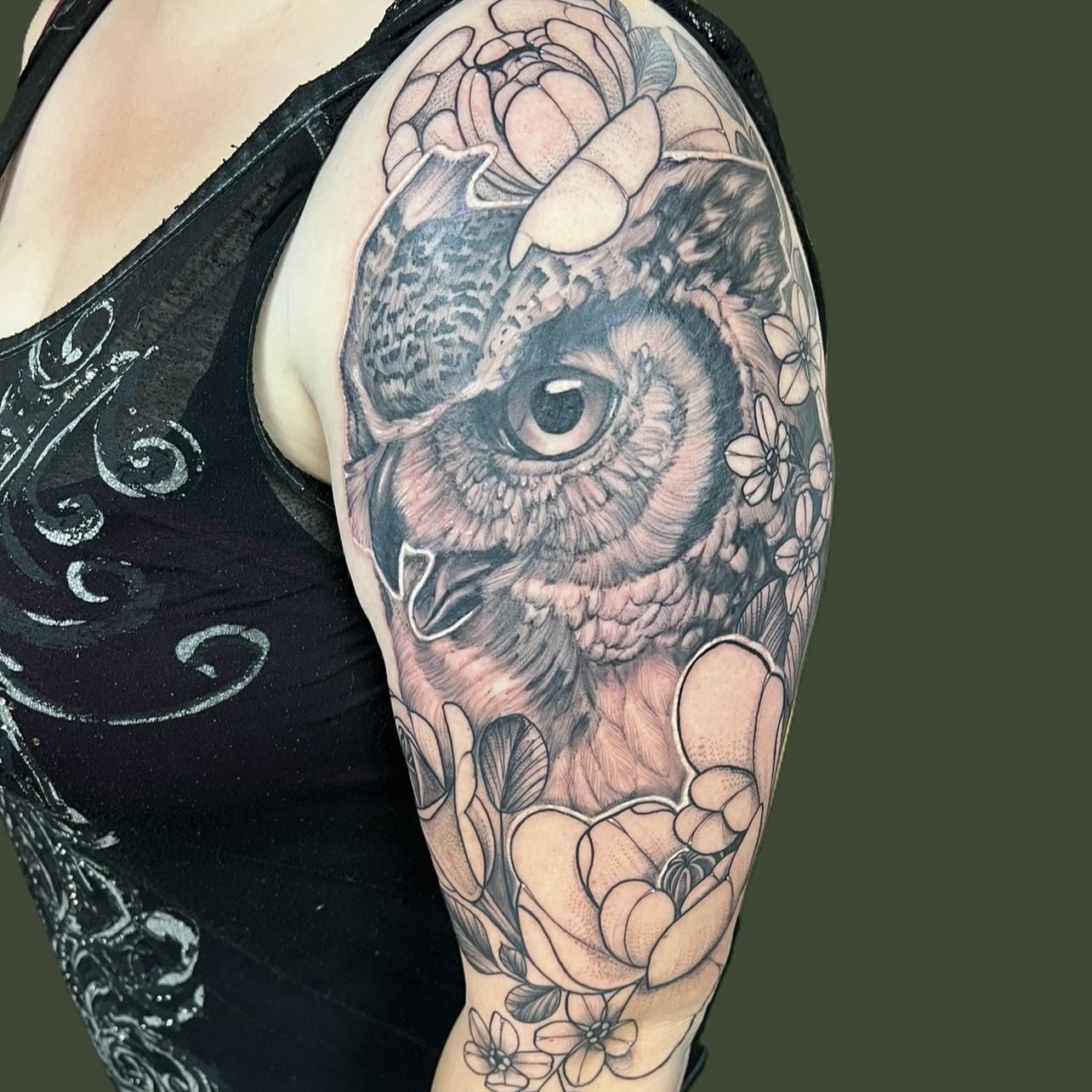 Black and grey tattoo on an upper arm: a realistic owl’s face with a piercing eye, framed by line-work peonies and small blossoms.