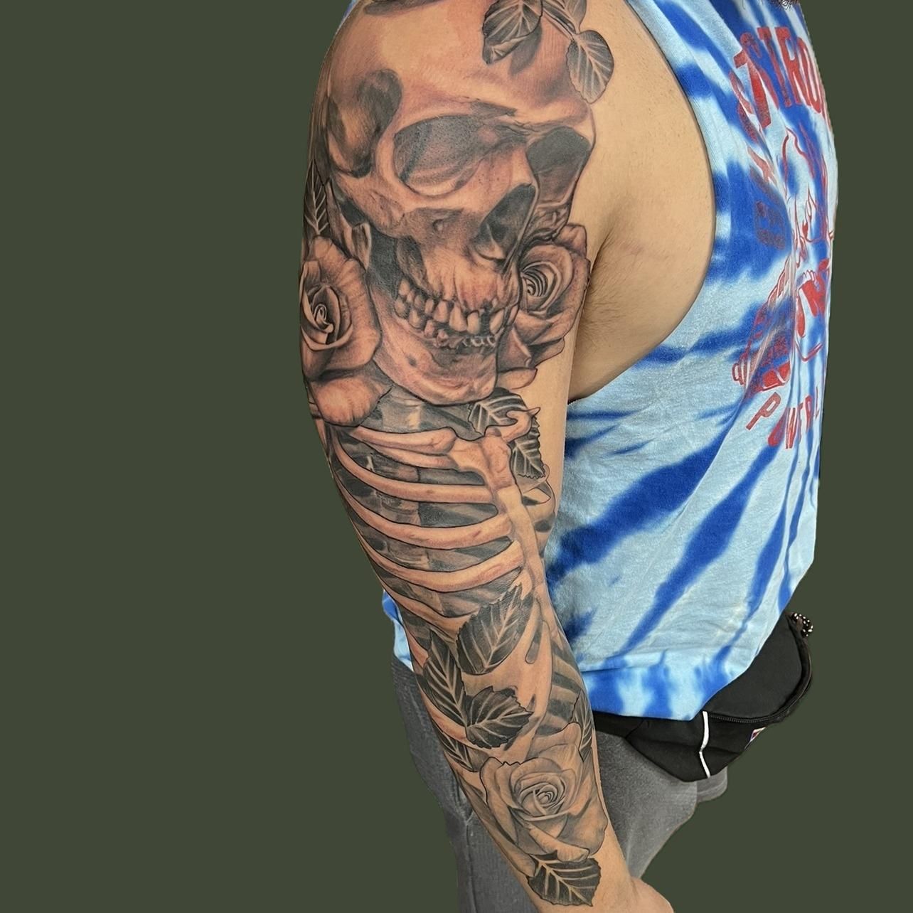 Black and grey sleeve tattoo: a realistic skull on the shoulder flanked by roses, flowing into a skeletal ribcage and leaves down the arm.