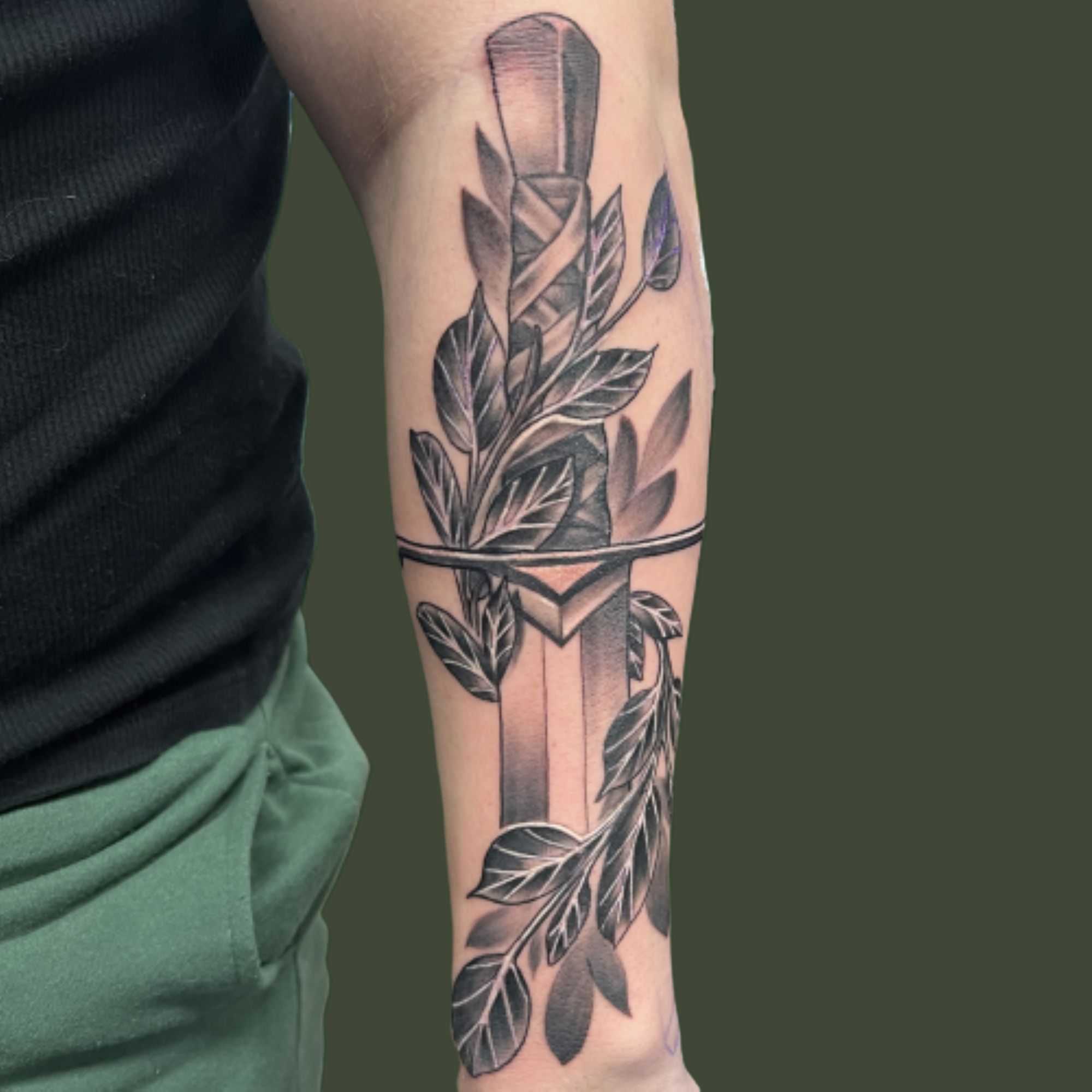 Black and grey forearm tattoo of a wrapped-hilt dagger entwined with shaded leafy branches, crossed by a thin band.
