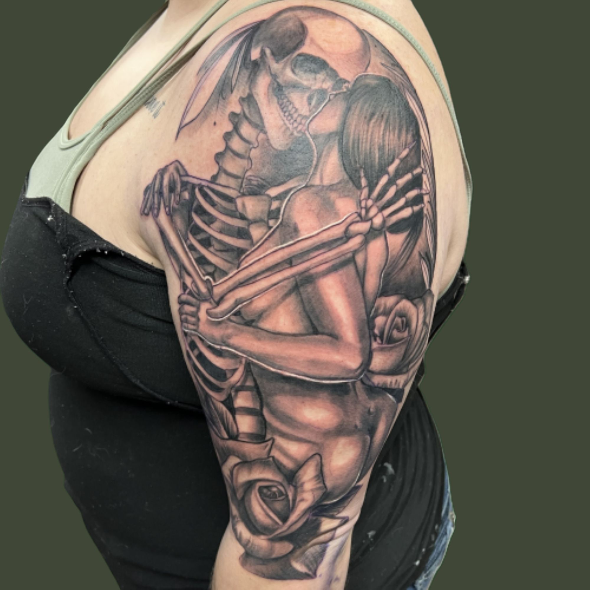 Black and grey upper-arm tattoo of a skeleton embracing and kissing a woman, with roses below and a pale moon behind.