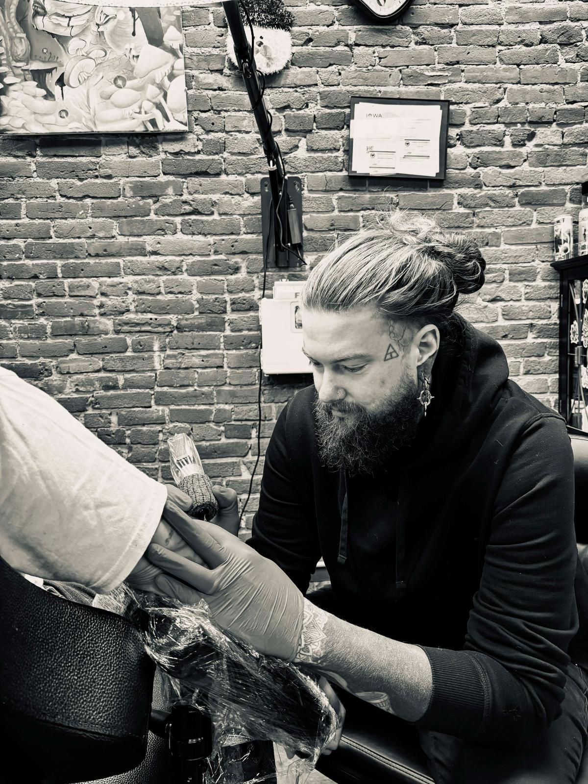 Black-and-white photo of Josh Clicquennoi, bearded with his hair tied back, tattooing a client’s arm in front of a brick wall.